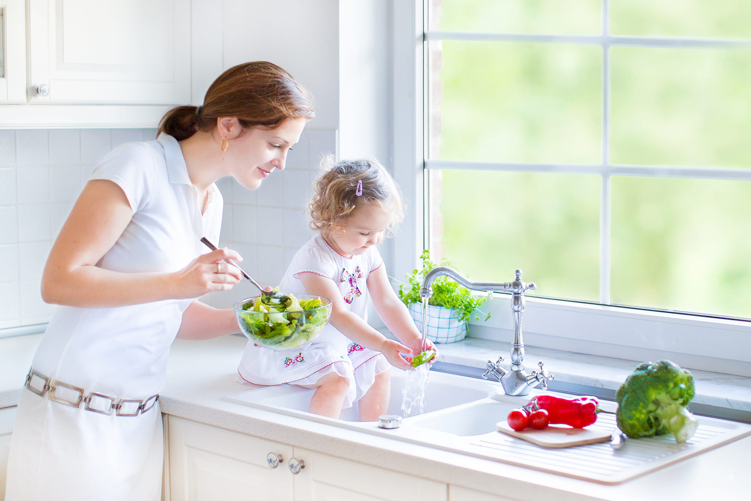 Criança sentada na pia lavando vegetais com sua mãe