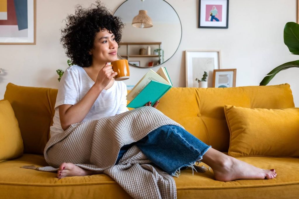 Mulher sentada no sofá lendo um livro e tomando um café