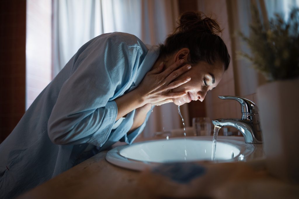 Mulher lavando o rosto em uma pia de banheiro com torneira tradicional