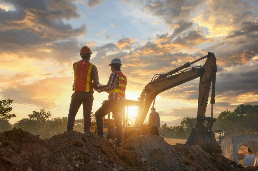 Dois homens profissionais da área de construção civil utilizando coletes na cor laranja, capacete, em cima de alguns montes de terra, ao fundo máquina escavadeira segurando um ART