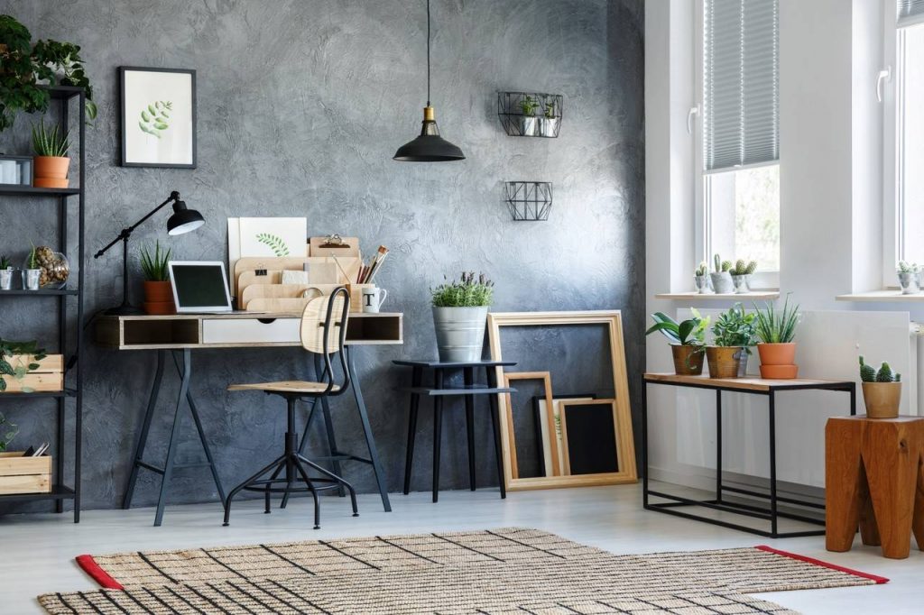 ambiente interno decorado com paredes de cimento queimado, escrivaninha com notebook e objetos sobrepostos, cadeira de escritório, canteiros com vaso de plantas sobrepostos.