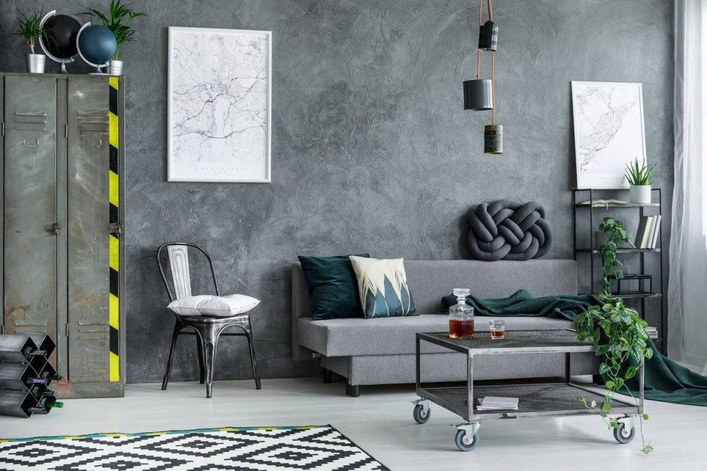 Sala  com paredes de cimento queimado, dois quadros decorativos de cor predominante branca fixos na parede, sofá no tom cinza com almofadas, mesa de centro em metal e de rodinhas.