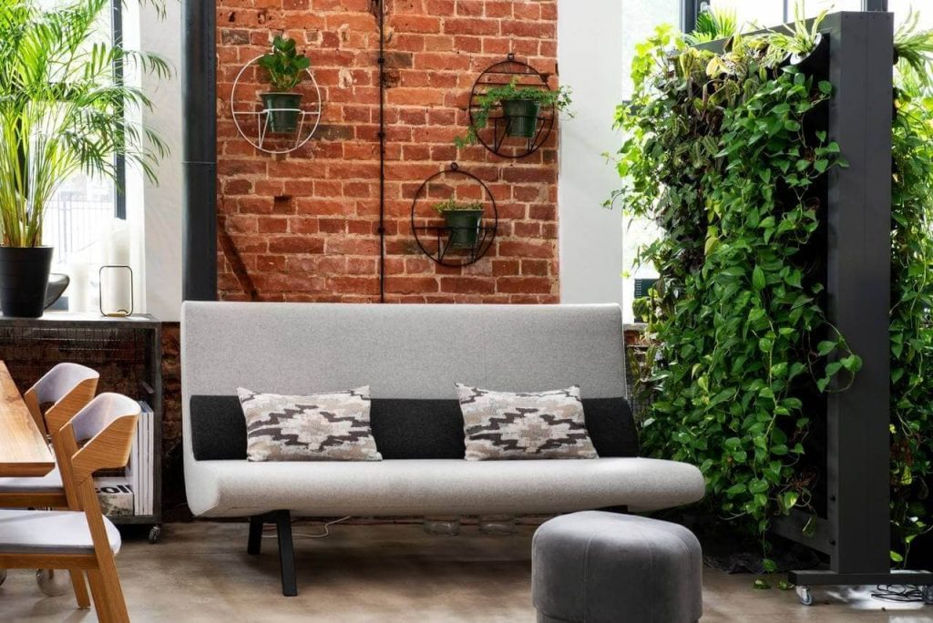 Ambiente interno com sofá no tom cinza, almofadas, puff, três vasos de plantas fixados na parede e jardim vertical