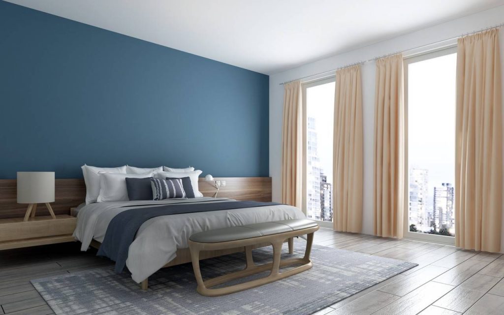 Dormitório com parede azul e branca, com cama de travesseiros brancos e colha cinza, ao pé o recamier almofadado no tom cinza
