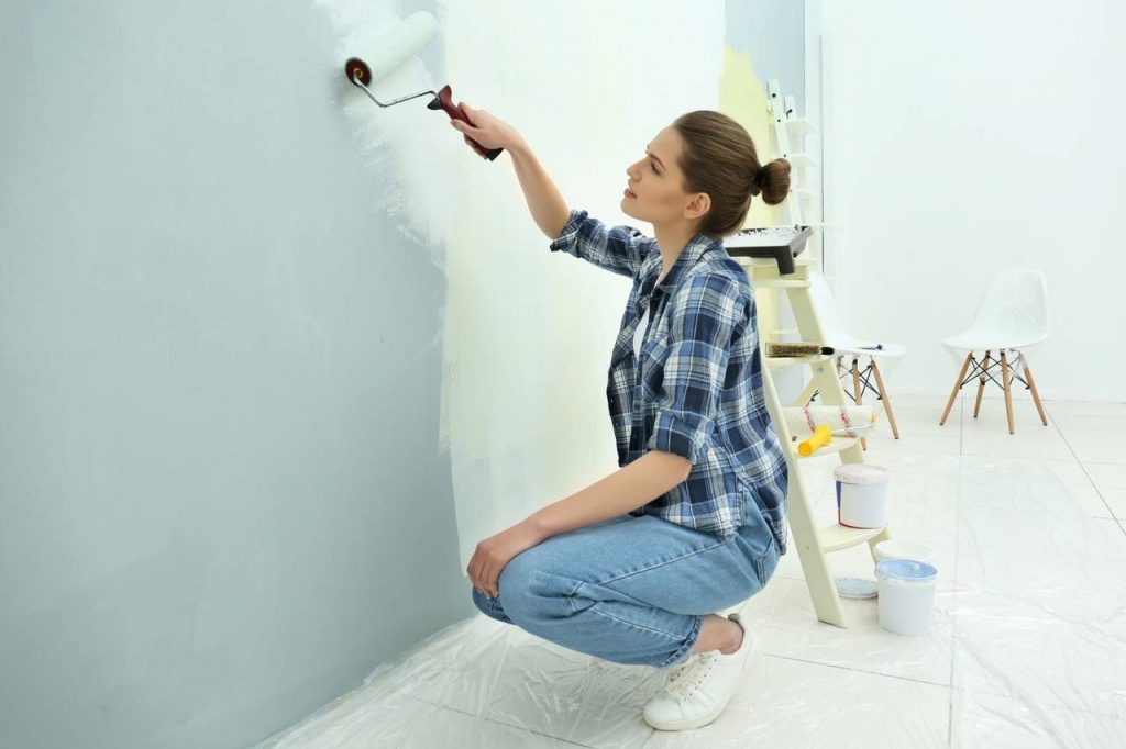 Mulher agachada realizando pintura na parede interna, ao lado latas de tinta e cadeiras