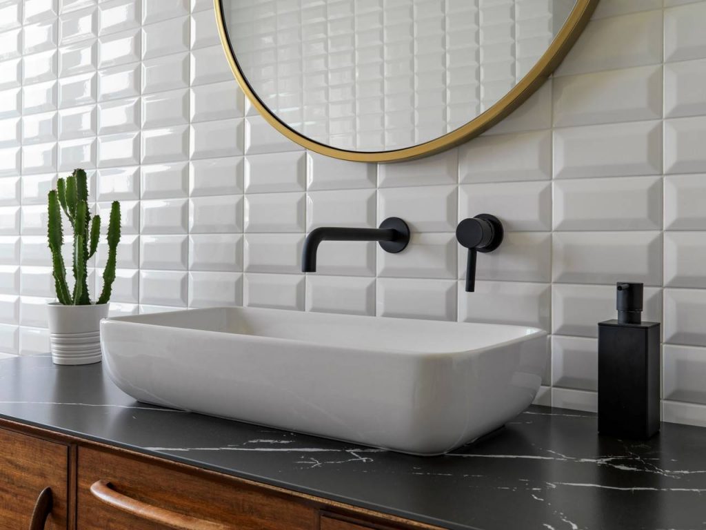Revestimento de parede de lavabo com porcelanato 3D na cor branca, espelho redondo fixo na parede, pia com cuba sobreposta, torneira de parede