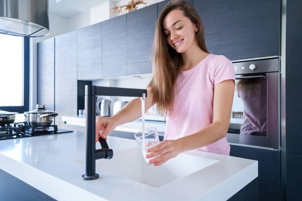 Mulher com Torneira 360º ligada e enchendo copo de vidro com água em cozinha