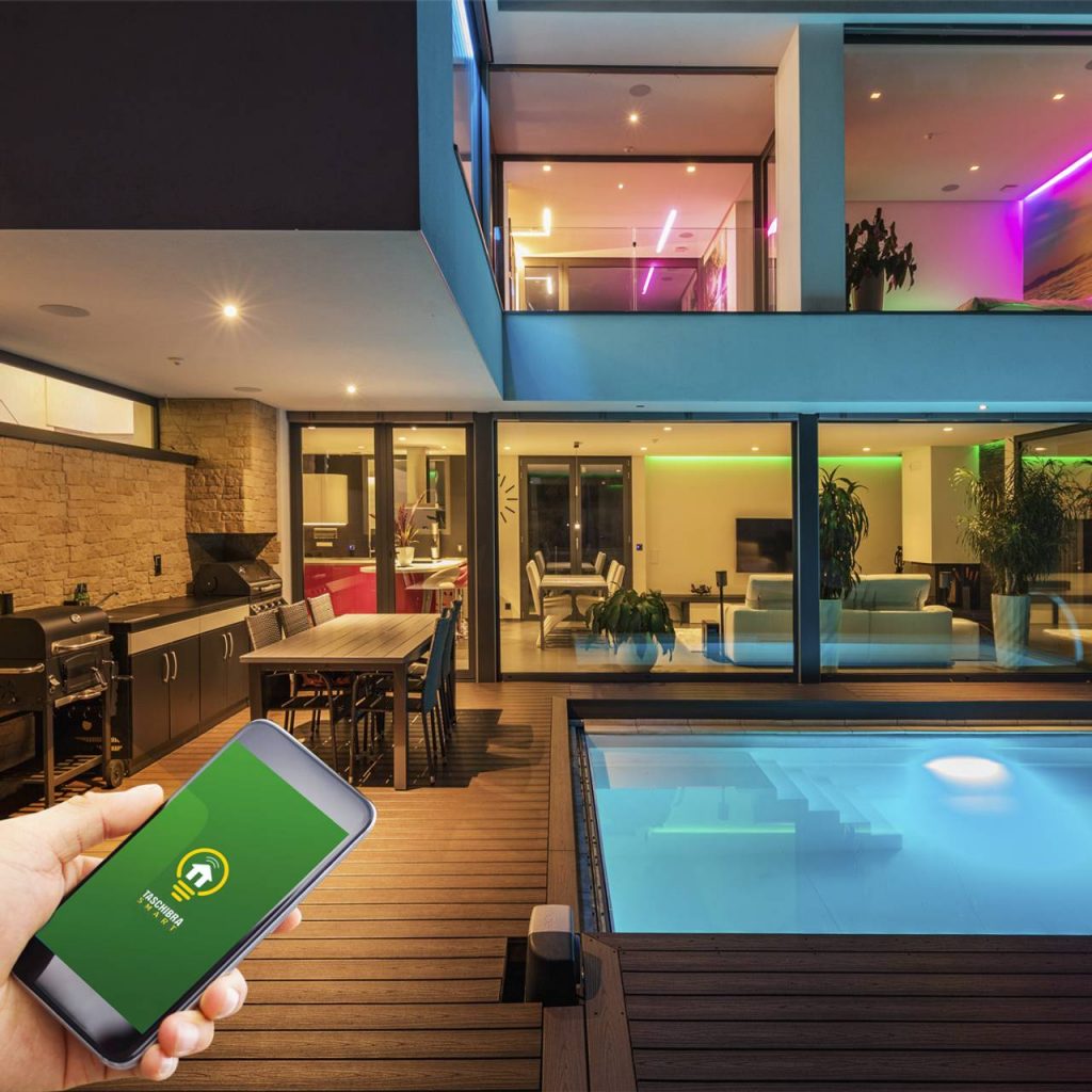 Detalhe de pessoa segurnado smartphone com aplicativo aberto de automação residencial, ao fundo casa com fachada em vidro, luzes coloridas e móveis