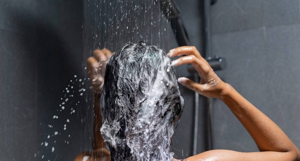 Detalhe de pessoa lavando cabelo cheio de espuma e com chuveiro ligado