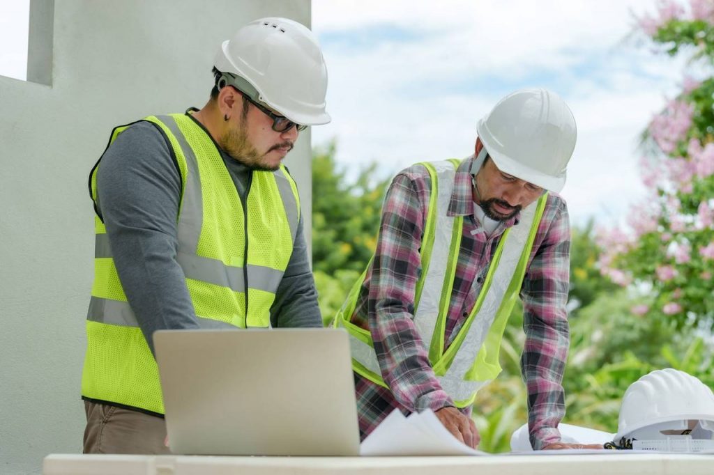 Dois trabalhadores com capacetes brancos olhando um projeto em cima da mesa utilizando epi