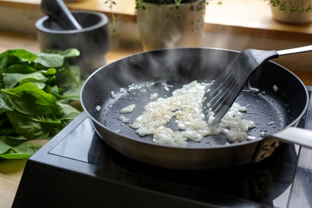 Detalhe de frigideira de cor preta fritando alho e com pessoa usando colher de silicone, que é um utensílio de cozinha, para mexer